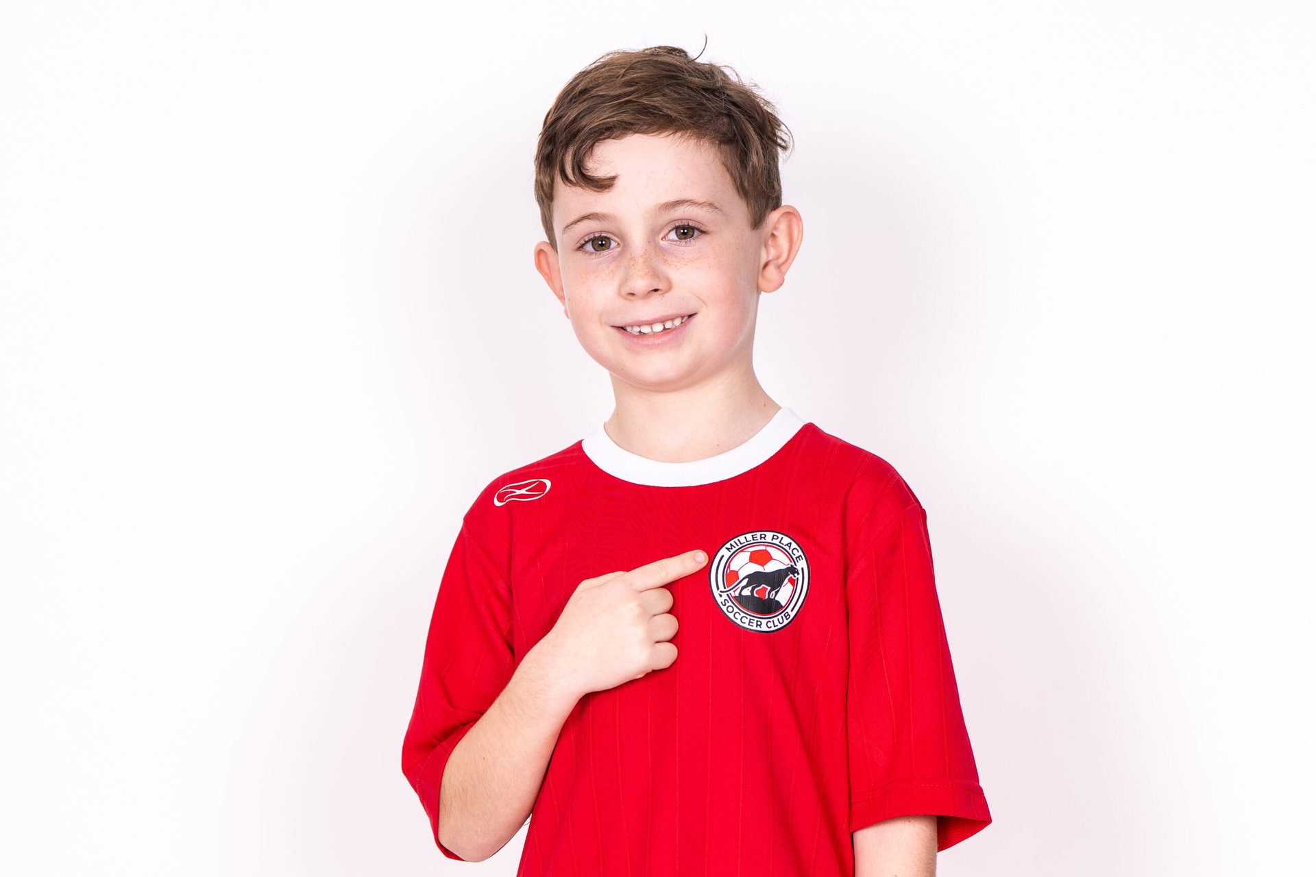 A young Miller Place Soccer Club player in a red jersey pointing to the club crest